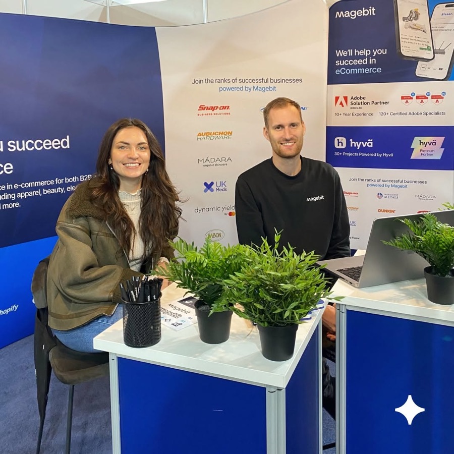 Two Magebit team members at the company's conference stand, with a wall of client logos behind them